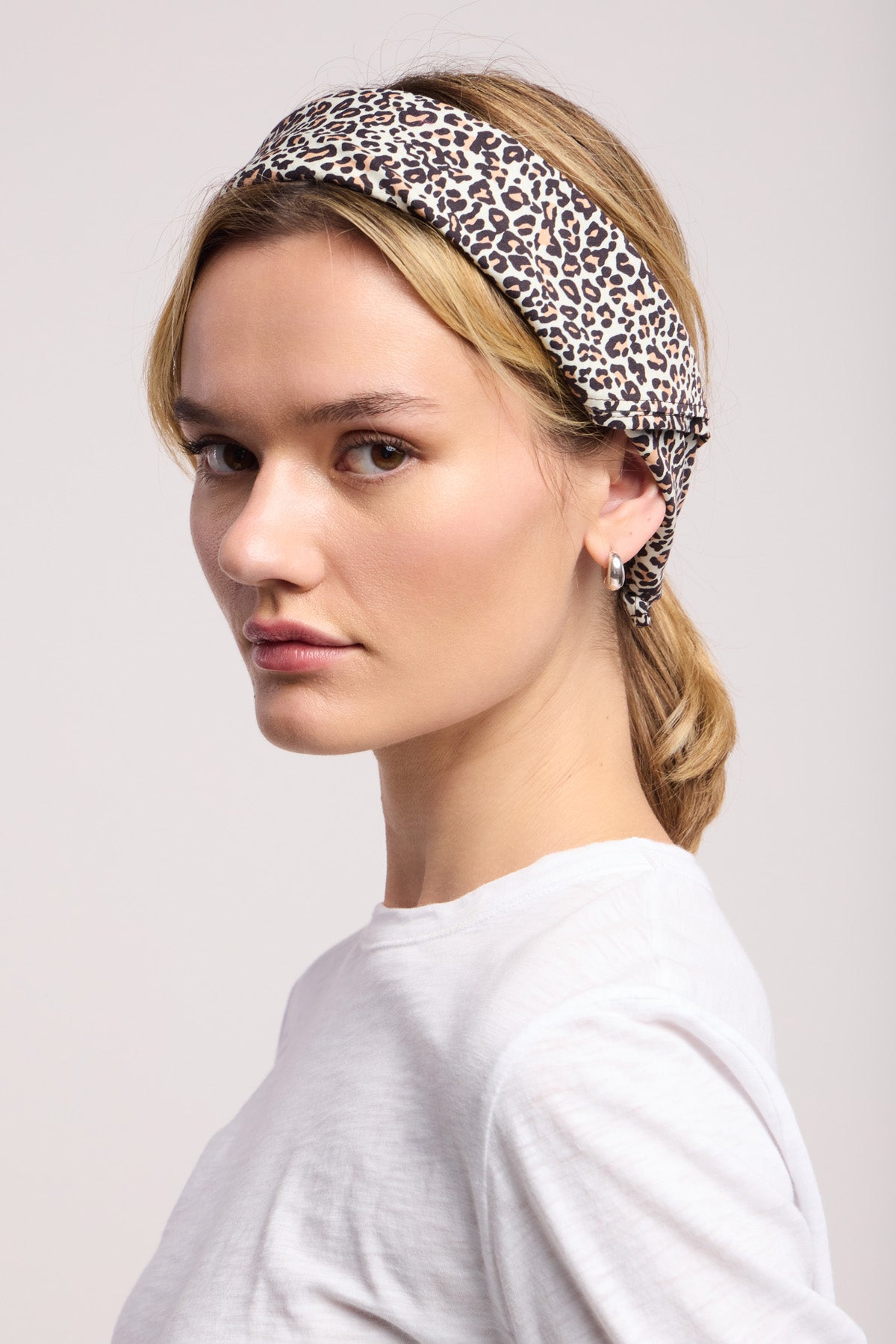 A woman wearing a white shirt and the JEWEL SCARF by Velvet by Graham & Spencer as a leopard print headband looks to the side against a plain light background.