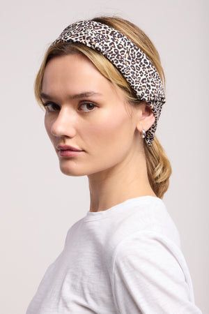 A woman wearing a white shirt and the JEWEL SCARF by Velvet by Graham & Spencer as a leopard print headband looks to the side against a plain light background.