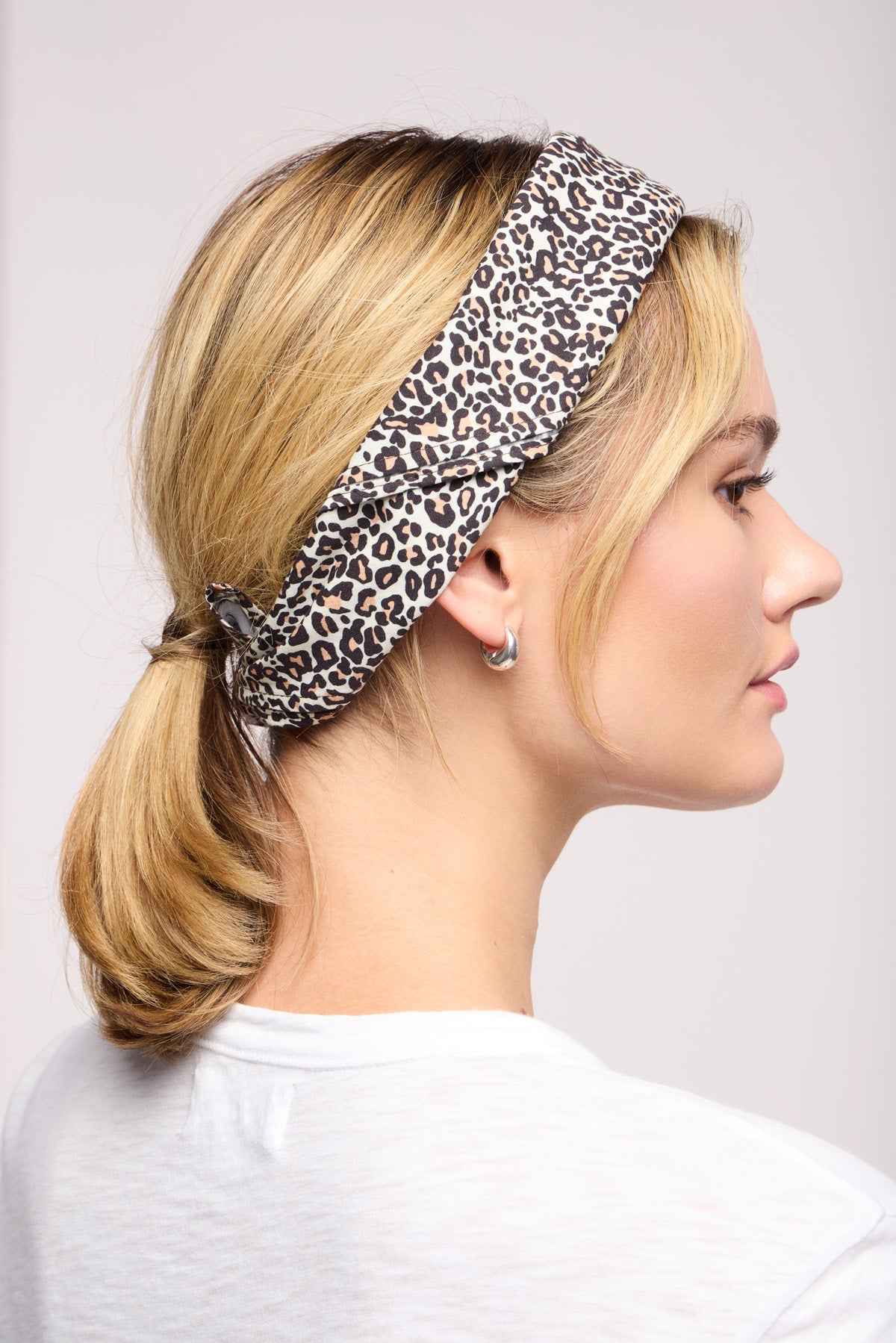 A woman with blonde hair wears the Velvet by Graham & Spencer JEWEL SCARF as a trendy headband and silver hoop earrings, shown in profile against a plain background.