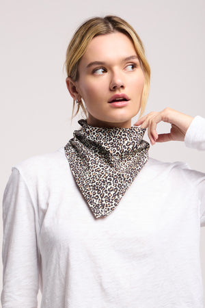A woman wears the JEWEL SCARF by Velvet by Graham & Spencer—a leopard print accessory—around her neck over a white long-sleeve shirt, looking to the side against a plain background.