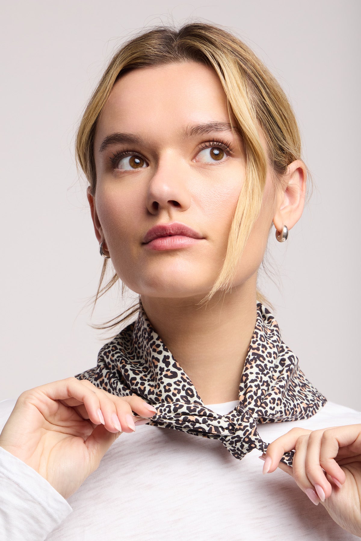 A woman with light hair in a white top adjusts the JEWEL SCARF by Velvet by Graham & Spencer, a lightweight, leopard print accessory, as she styles this versatile scarf around her neck and looks slightly upward.-40453022351553