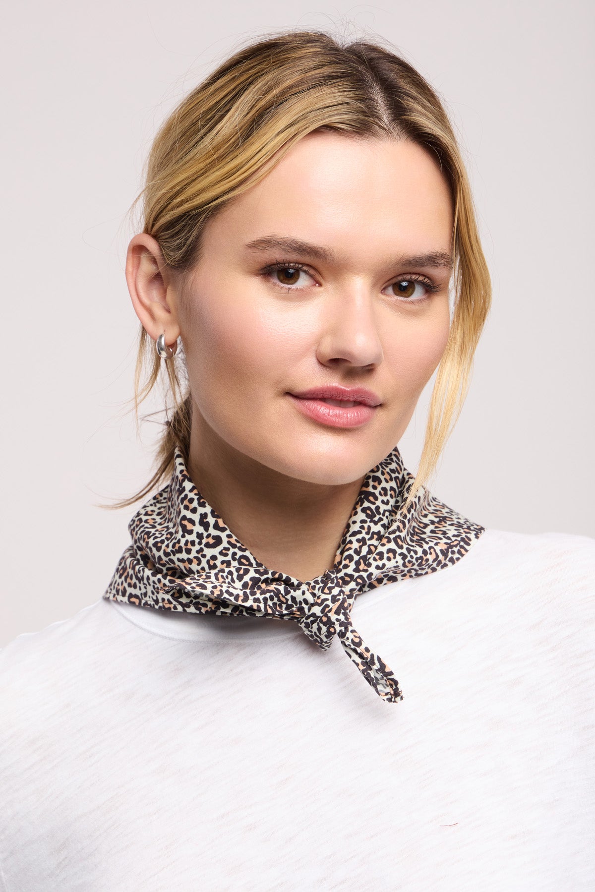 A woman with light hair wears a white top and the JEWEL SCARF by Velvet by Graham & Spencer, featuring a leopard print, around her neck while looking at the camera with a neutral expression.-40453022318785