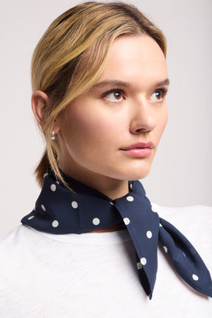 A person with light hair wears the JEWEL SCARF by Velvet by Graham & Spencer—a lightweight navy blue scarf with white polka dots—around their neck and a white top, looking slightly to the side.