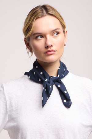 A woman wearing a white shirt and the JEWEL SCARF by Velvet by Graham & Spencer—navy blue with polka dots—tied at her neck, looks slightly to the side against a plain background.