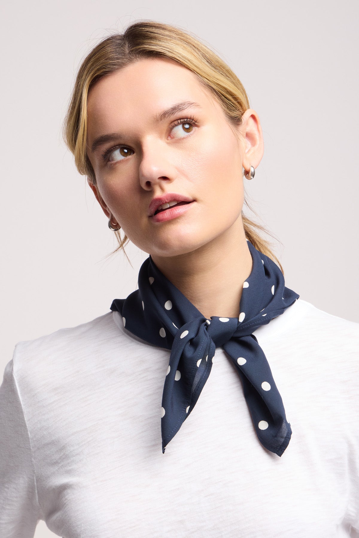 A person wearing a white shirt and the JEWEL SCARF by Velvet by Graham & Spencer—a lightweight navy blue and white polka dot scarf—tied around their neck, looking upwards.