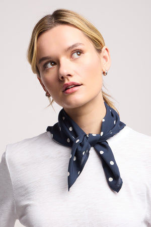 A person wearing a white shirt and the JEWEL SCARF by Velvet by Graham & Spencer—a lightweight navy blue and white polka dot scarf—tied around their neck, looking upwards.