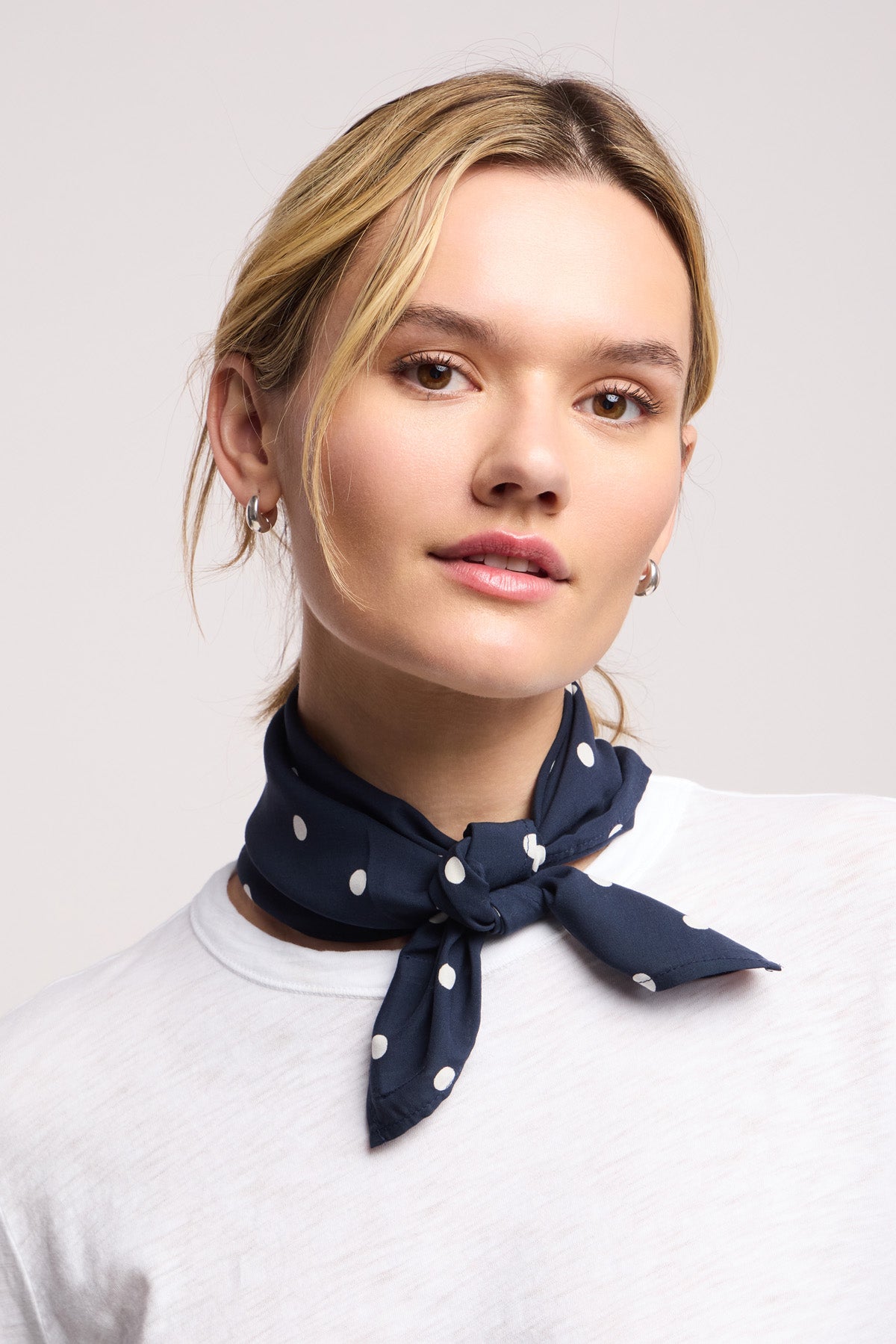 A person wearing a white shirt and the JEWEL SCARF by Velvet by Graham & Spencer—navy blue with white polka dots—tied around their neck, light hair pulled back, and small hoop earrings.