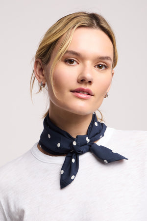 A person wearing a white shirt and the JEWEL SCARF by Velvet by Graham & Spencer—navy blue with white polka dots—tied around their neck, light hair pulled back, and small hoop earrings.