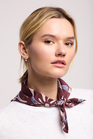 A woman with blonde hair models the Velvet by Graham & Spencer JEWEL SCARF in a maroon pattern, styled around her neck over a white top, against a plain background.
