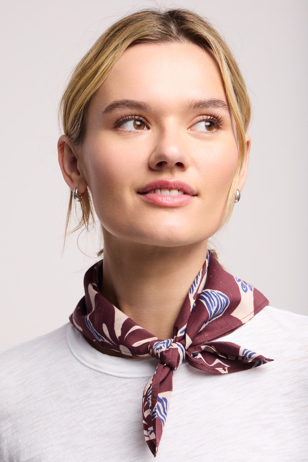 A woman with blonde hair tied back wears a white top and the JEWEL SCARF by Velvet by Graham & Spencer, a versatile maroon accessory loosely knotted around her neck as she gazes slightly upward and to the side.-40453004787905
