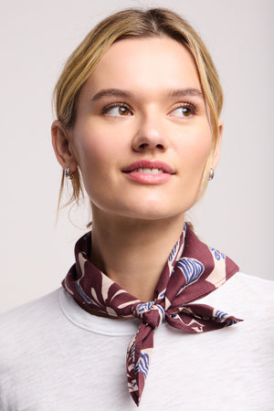A woman with blonde hair tied back wears a white top and the JEWEL SCARF by Velvet by Graham & Spencer, a versatile maroon accessory loosely knotted around her neck as she gazes slightly upward and to the side.