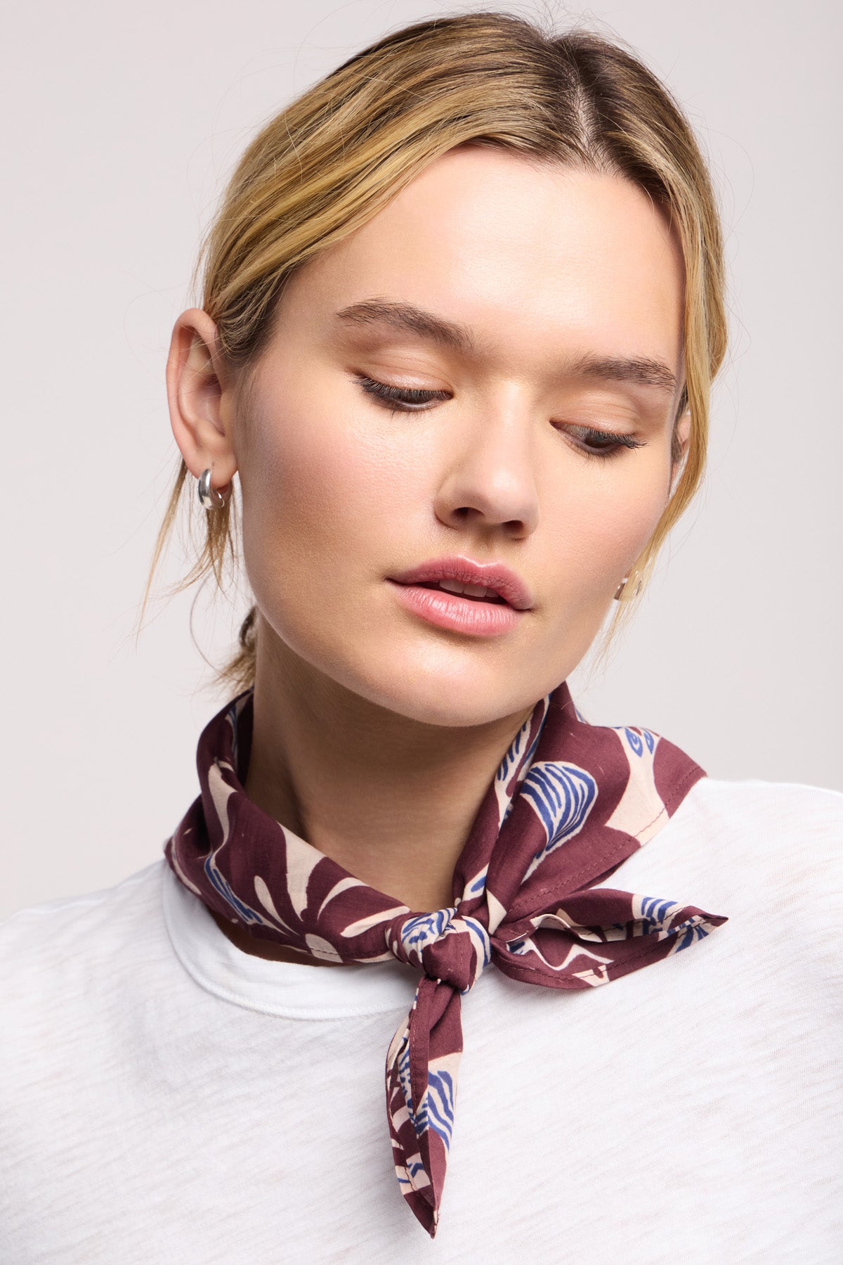 A woman with blond hair wears a white shirt and the Velvet by Graham & Spencer JEWEL SCARF in brown, tied around her neck, looking slightly downward with a neutral expression.-40453004722369