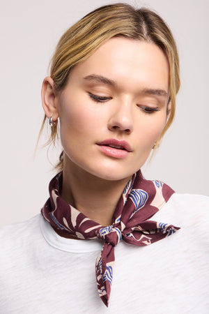A woman with blond hair wears a white shirt and the Velvet by Graham & Spencer JEWEL SCARF in brown, tied around her neck, looking slightly downward with a neutral expression.
