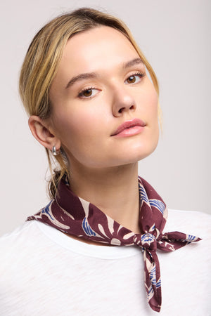 A woman with blonde hair tied back wears a white top and the JEWEL SCARF by Velvet by Graham & Spencer, a lightweight maroon scarf with blue and beige patterns, tied around her neck.