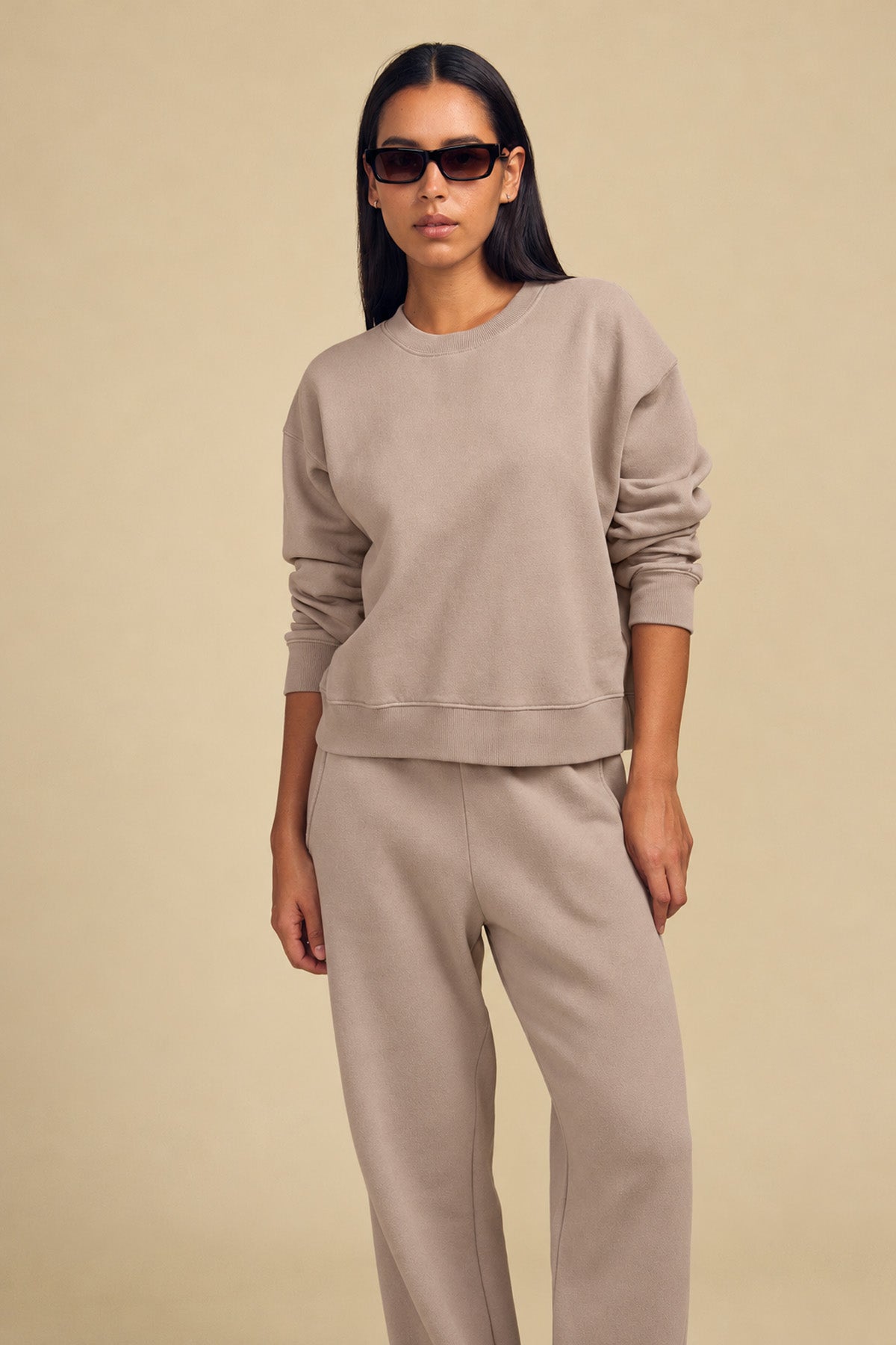 A person wearing a tan YOKO SWEATSHIRT and matching relaxed-fit cotton fleece pants, styled with dark sunglasses.