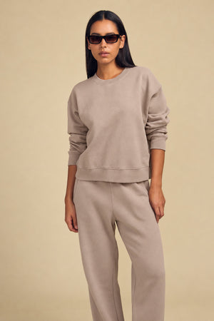 A person wearing a tan YOKO SWEATSHIRT and matching relaxed-fit cotton fleece pants, styled with dark sunglasses.