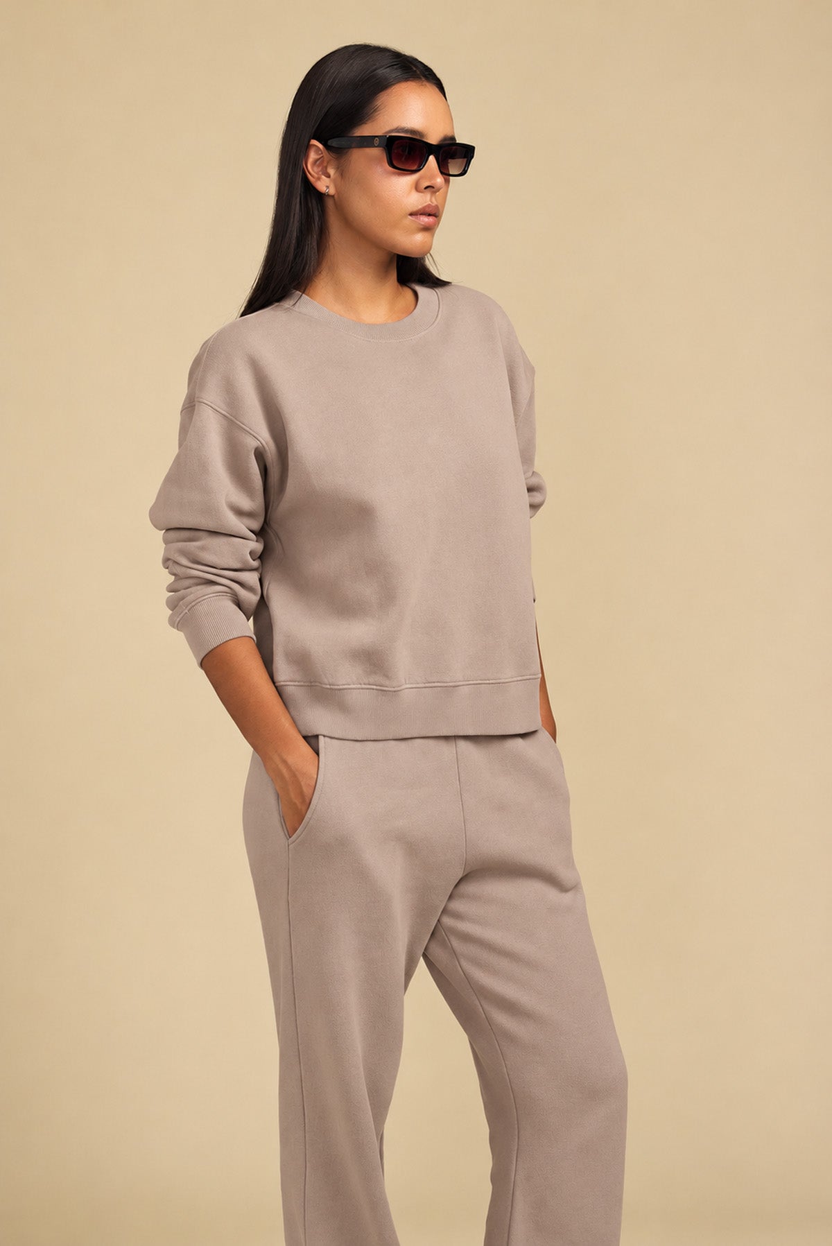 A person wearing a relaxed YOKO SWEATSHIRT and matching taupe sweatpants with pockets, and dark sunglasses.-46320580296897