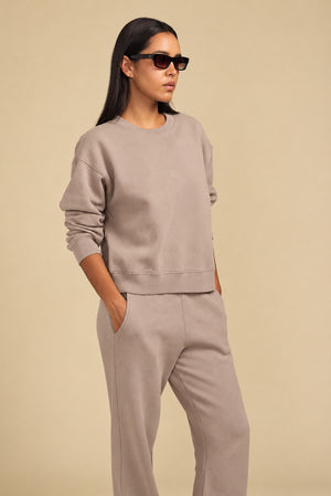 A person wearing a relaxed YOKO SWEATSHIRT and matching taupe sweatpants with pockets, and dark sunglasses.