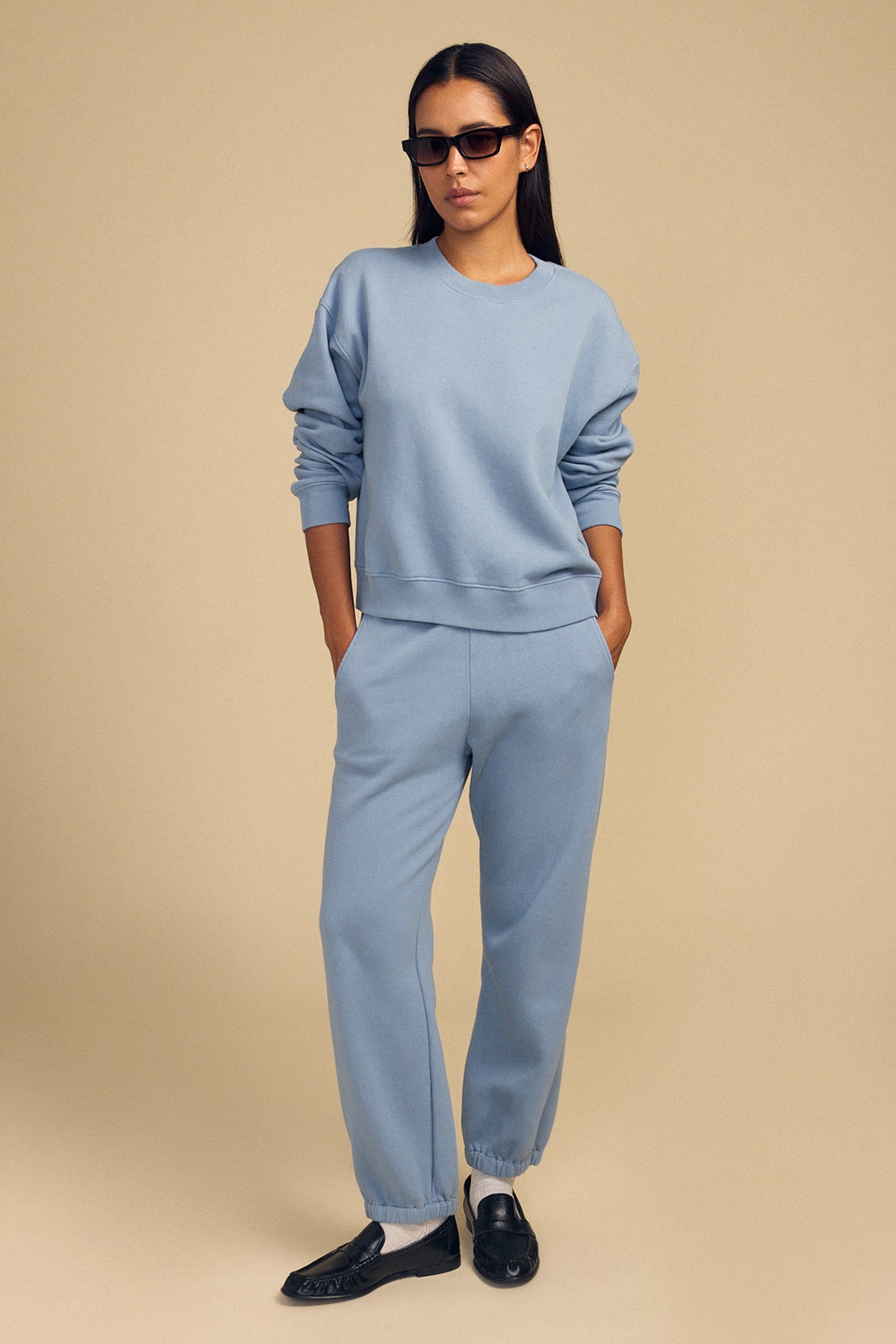 A woman wears black sunglasses, a light blue sweatshirt, and Velvet by Graham & Spencer ZARA SWEATPANT, standing with hands in pockets against a beige background.