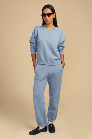 A woman wears black sunglasses, a light blue sweatshirt, and Velvet by Graham & Spencer ZARA SWEATPANT, standing with hands in pockets against a beige background.