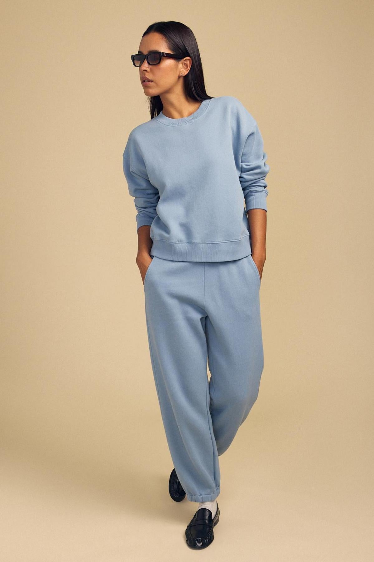 Woman wearing sunglasses, a light blue cotton fleece sweatshirt, and ZARA SWEATPANT by Velvet by Graham & Spencer stands with hands in pockets against a beige background.