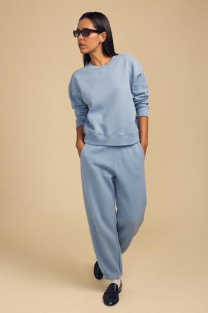 Woman wearing sunglasses, a light blue cotton fleece sweatshirt, and ZARA SWEATPANT by Velvet by Graham & Spencer stands with hands in pockets against a beige background.