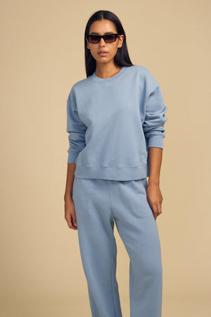 A person in sunglasses and a light blue ZARA SWEATPANT by Velvet by Graham & Spencer stands against a beige background, highlighting the relaxed fit in soft cotton fleece.