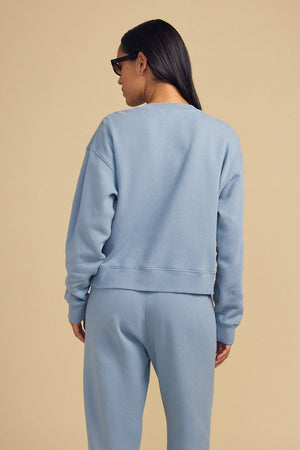 Person in a light blue cotton fleece sweatshirt and ZARA SWEATPANT by Velvet by Graham & Spencer stands facing away against a beige background, wearing sunglasses.