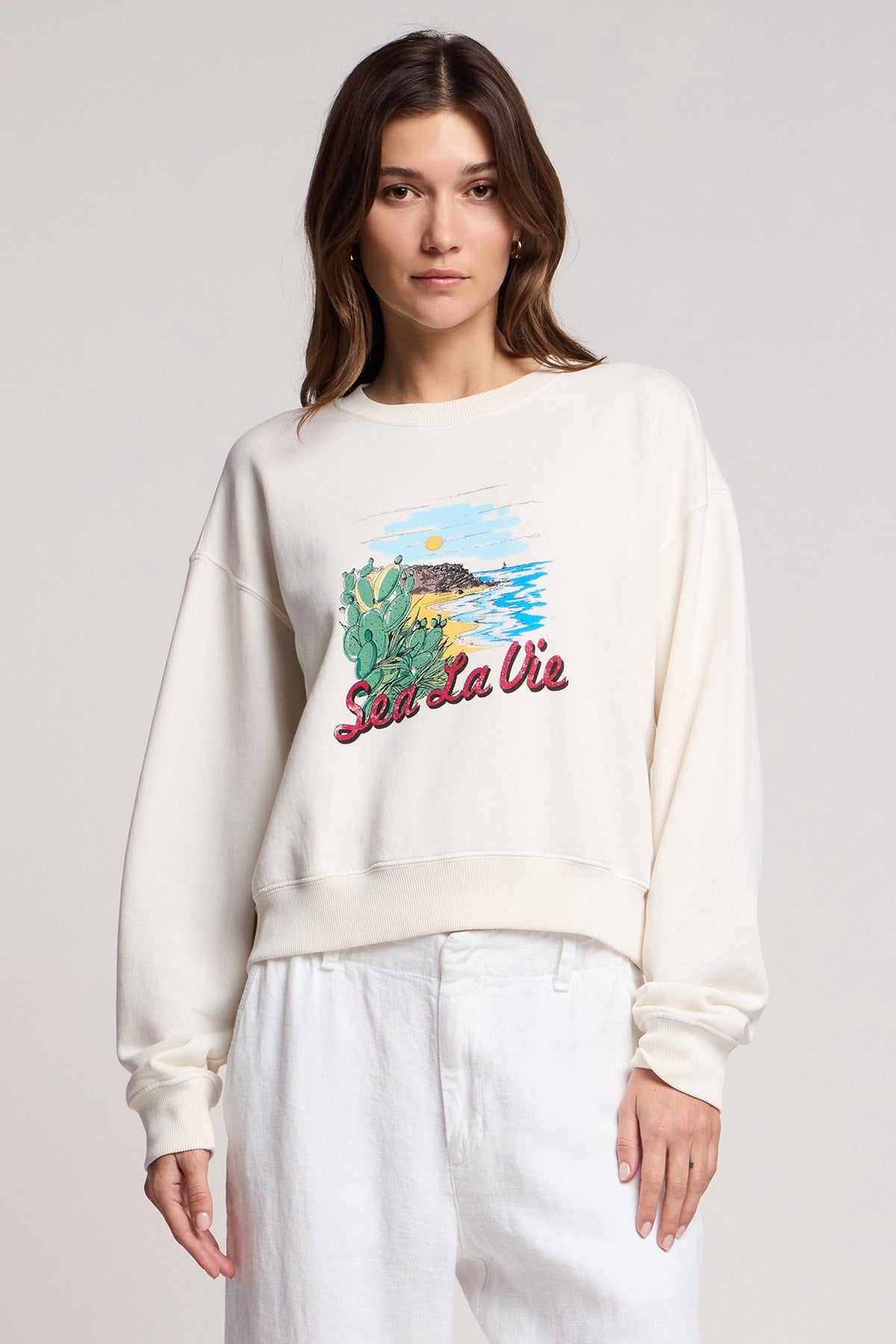 A woman wears the KENNA SWEATSHIRT by Velvet by Graham & Spencer, a cream-colored cotton fleece with a California coast scene and 