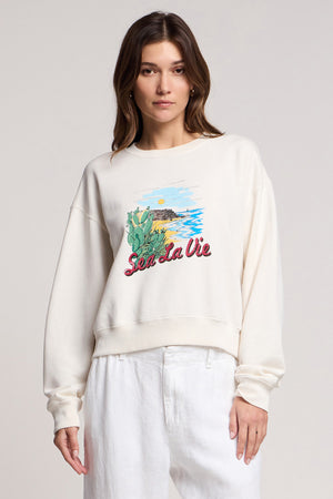 A woman wears the KENNA SWEATSHIRT by Velvet by Graham & Spencer, a cream-colored cotton fleece with a California coast scene and 