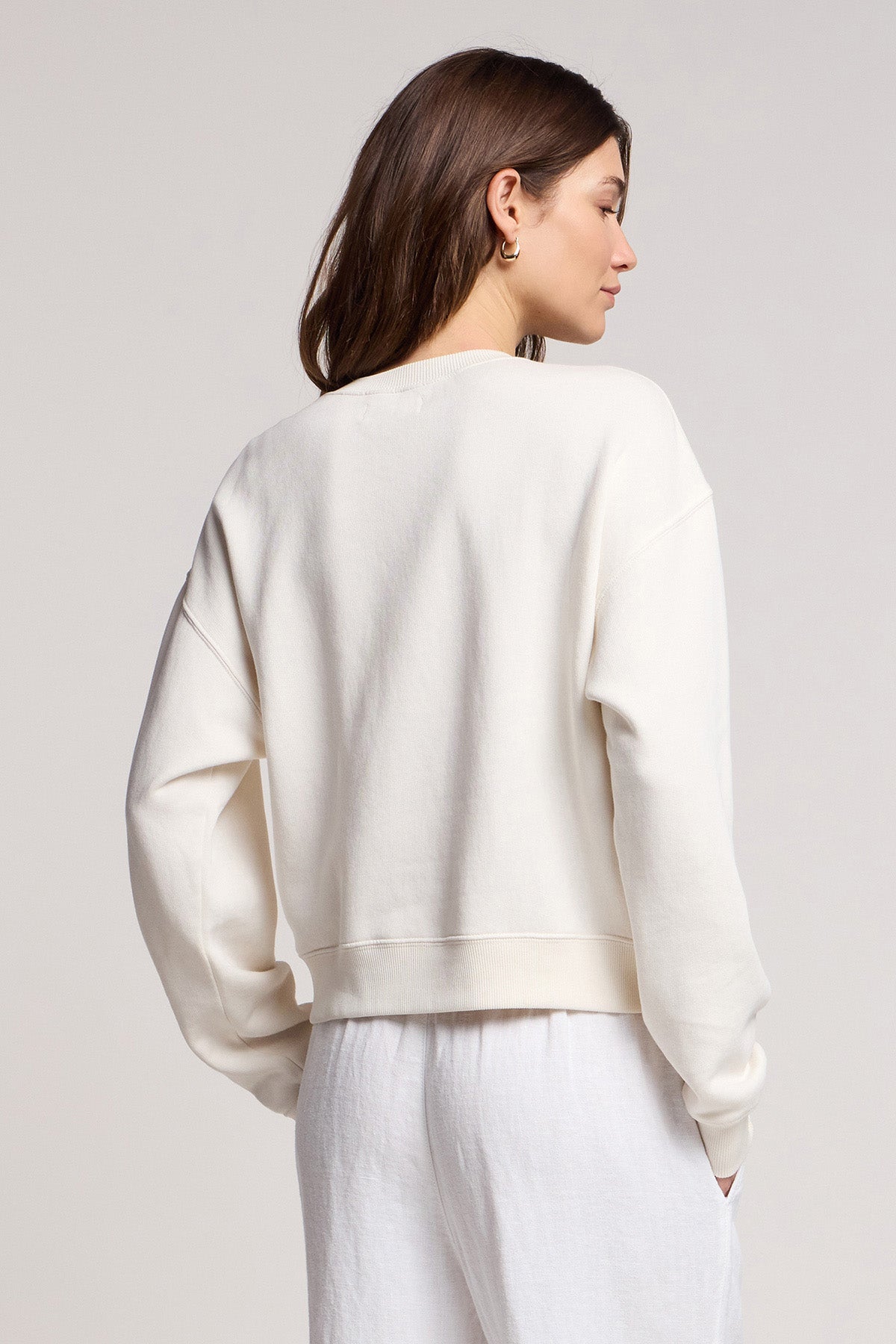 A person with long brown hair wears the off-white KENNA SWEATSHIRT by Velvet by Graham & Spencer and white pants, photographed from behind against a light background.