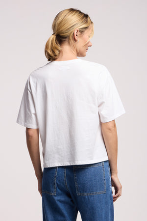 A person stands with back to camera, wearing the SAMMY TEE by Velvet by Graham & Spencer in white cotton jersey and blue jeans, set against a plain background.