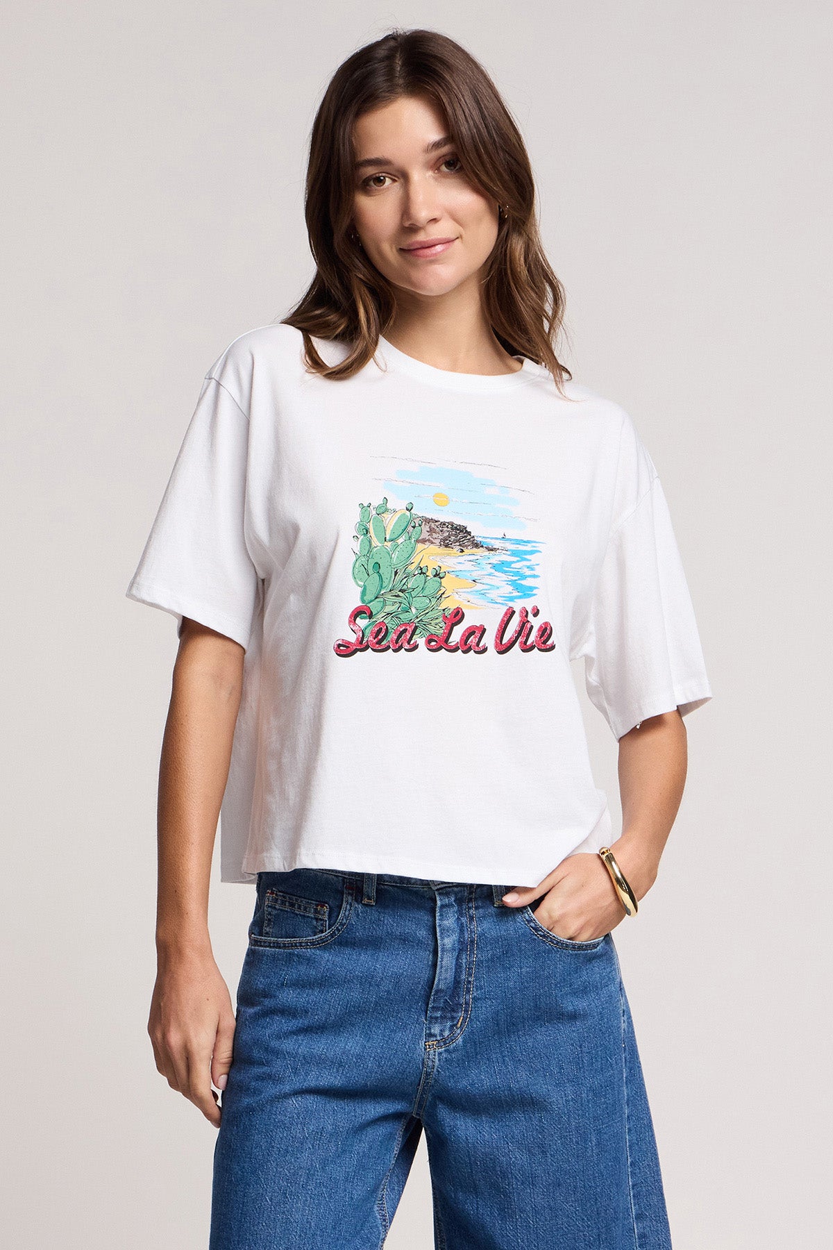 A woman wears the SAMMY TEE by Velvet by Graham & Spencer—a soft cotton jersey top with a vibrant "Sea La Vie" graphic—paired with blue jeans, standing against a simple light background.-41372208201921