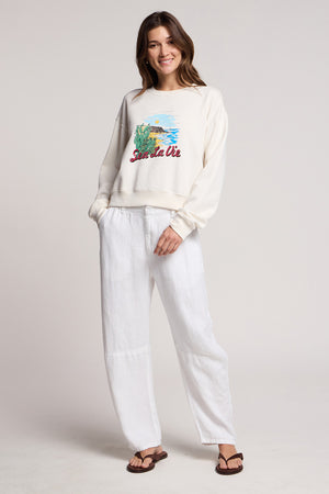 A woman faces forward wearing the KENNA SWEATSHIRT by Velvet by Graham & Spencer—a cream cotton fleece graphic top inspired by the California coast—paired with loose white pants and brown sandals against a plain background.