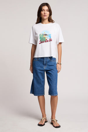 A woman stands against a plain background wearing the white SAMMY TEE from Velvet by Graham & Spencer, paired with knee-length denim shorts and black flip-flops.
