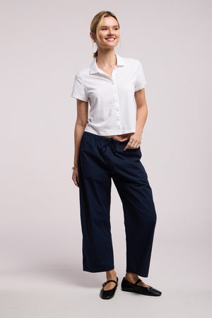 A person wearing the Velvet by Graham & Spencer ARLENE POLO, a short-sleeved white button-up with a relaxed boxy fit, navy wide-leg pants, and black flats stands smiling against a plain light background.