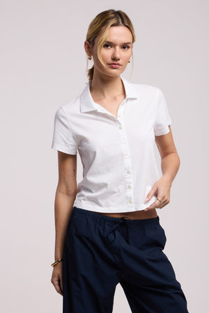 A person wearing the ARLENE POLO by Velvet by Graham & Spencer, a relaxed boxy white short-sleeve button-up shirt, and navy blue drawstring pants stands against a plain light background.