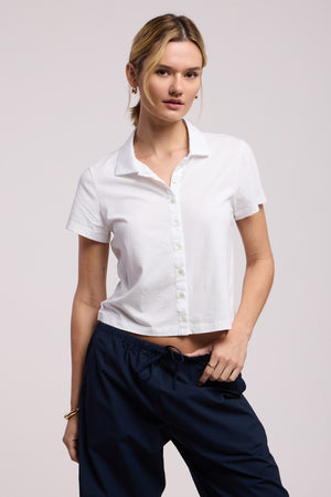 A person stands against a plain light background wearing the ARLENE POLO by Velvet by Graham & Spencer—a short-sleeved white button-up shirt with a relaxed, boxy silhouette—paired with dark blue drawstring pants.