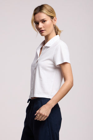 A woman stands sideways against a plain light background wearing the ARLENE POLO by Velvet by Graham & Spencer, a relaxed boxy short-sleeve white cotton jersey button-up shirt paired with dark pants.