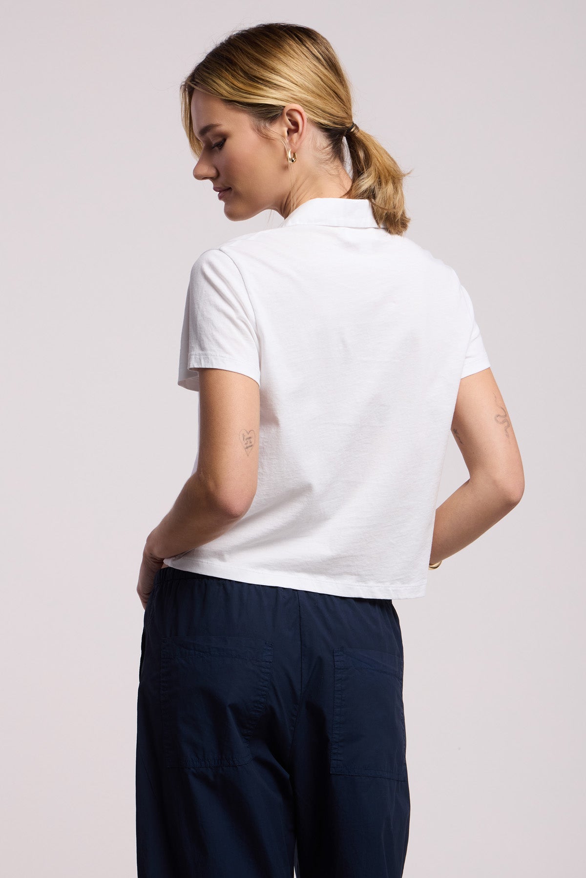 A person stands with their back to the camera, wearing the ARLENE POLO by Velvet by Graham & Spencer—a white short-sleeve polo shirt in a relaxed boxy fit—paired with dark blue pants and sporting a low ponytail.-41371452670145