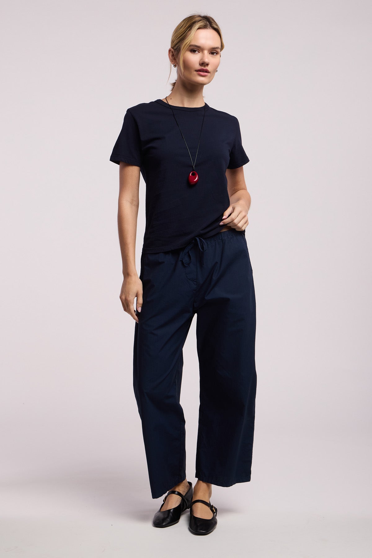A person wears the RYLAND TEE by Velvet by Graham & Spencer, paired with loose dark pants, black flats, and a long necklace with a red pendant against a plain light background.-41372113567937