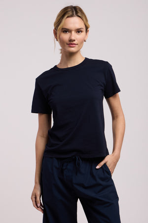 A person wears the RYLAND TEE by Velvet by Graham & Spencer— a plain black, boxy cotton jersey t-shirt— paired with dark pants, standing against a neutral background with one hand in their pocket.