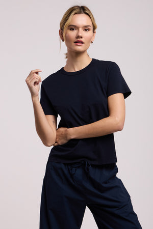 A person faces forward wearing the RYLAND TEE by Velvet by Graham & Spencer, a plain black cotton jersey top, with dark pants against a light neutral background.