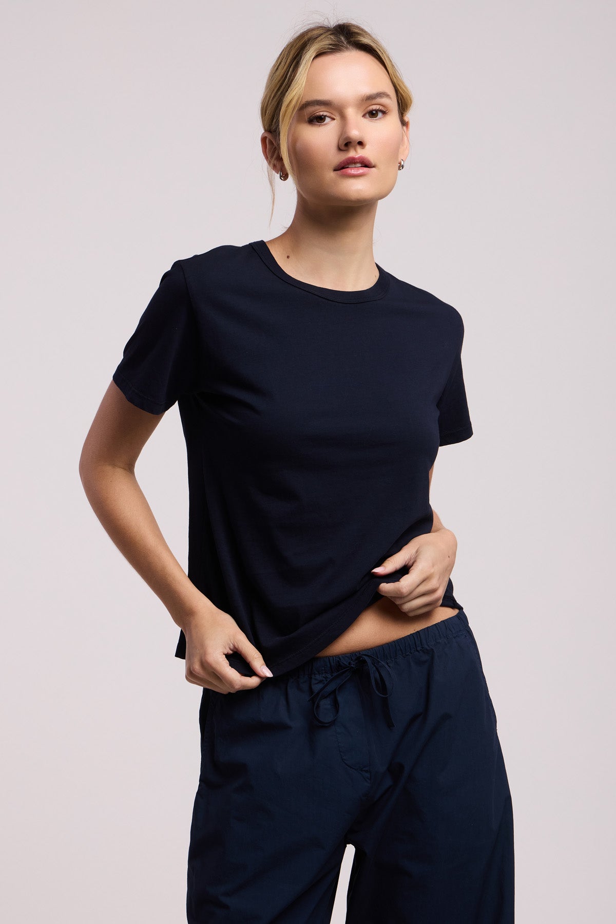 A person wearing the RYLAND TEE by Velvet by Graham & Spencer—a plain black tee with a boxy silhouette—and black drawstring pants stands against a neutral background, holding the hem of their shirt with one hand.