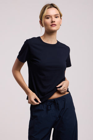 A person wearing the RYLAND TEE by Velvet by Graham & Spencer—a plain black tee with a boxy silhouette—and black drawstring pants stands against a neutral background, holding the hem of their shirt with one hand.