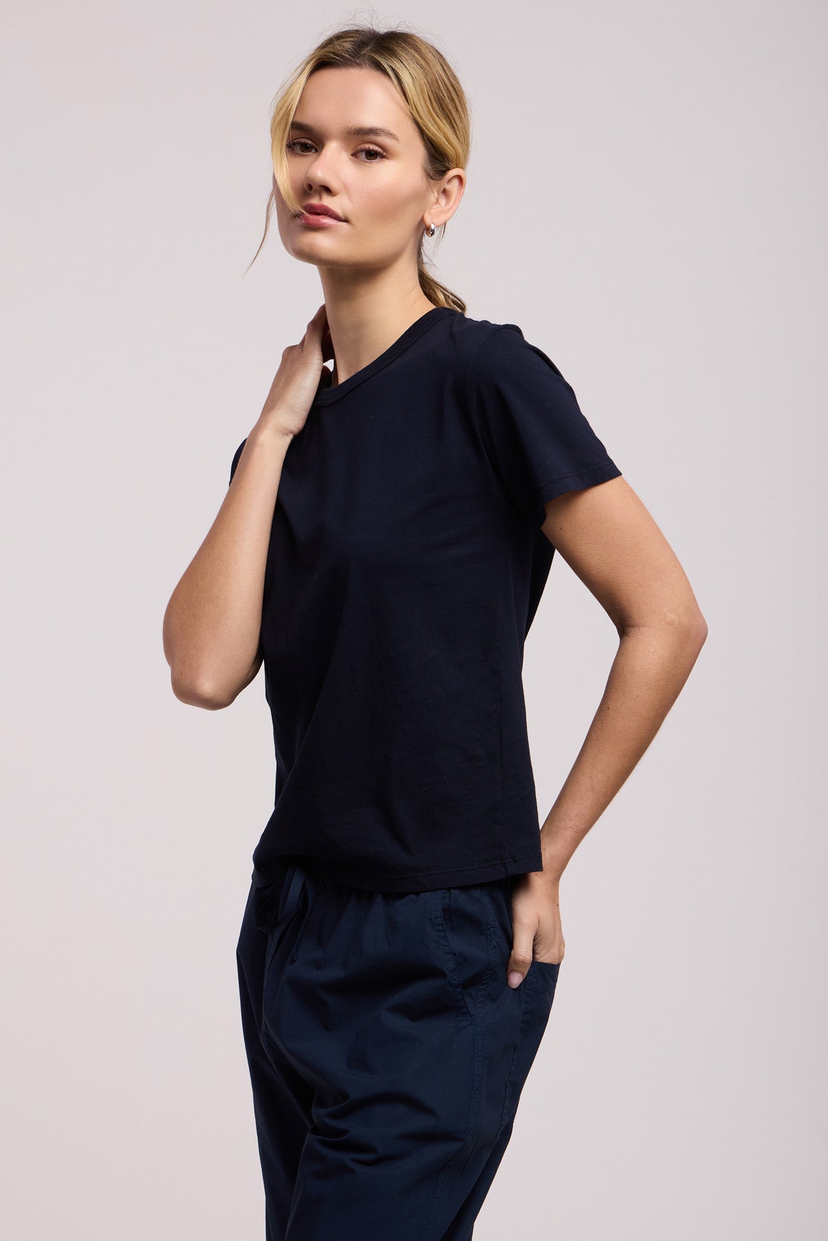 A woman wears the RYLAND TEE by Velvet by Graham & Spencer—a modern, boxy cotton jersey tee—paired with dark pants. She poses against a neutral background, one hand on her neck and the other in her pocket.