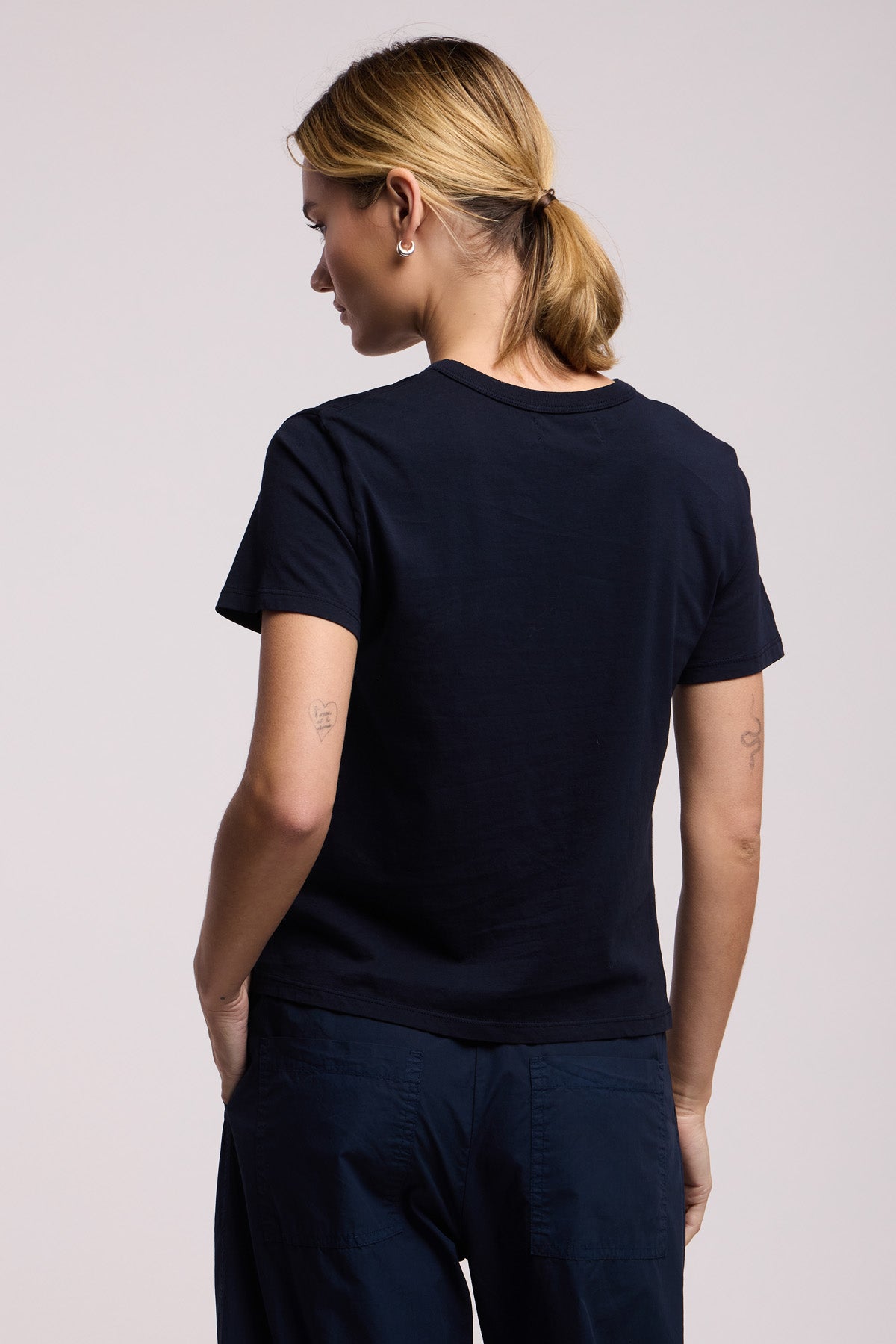 A person with blonde hair in a low ponytail is shown from the back wearing the RYLAND TEE by Velvet by Graham & Spencer, styled with dark pants and standing against a light background.-41372113895617
