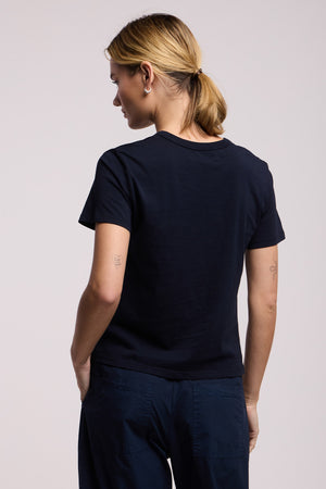 A person with blonde hair in a low ponytail is shown from the back wearing the RYLAND TEE by Velvet by Graham & Spencer, styled with dark pants and standing against a light background.