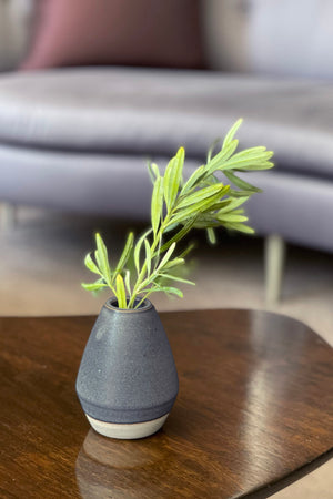 A CERAMIC BUD VASE from Jenny Graham Home with green leaves placed on a wooden table, with a blurred couch in the background, adds a touch of charm to the guest bathroom.