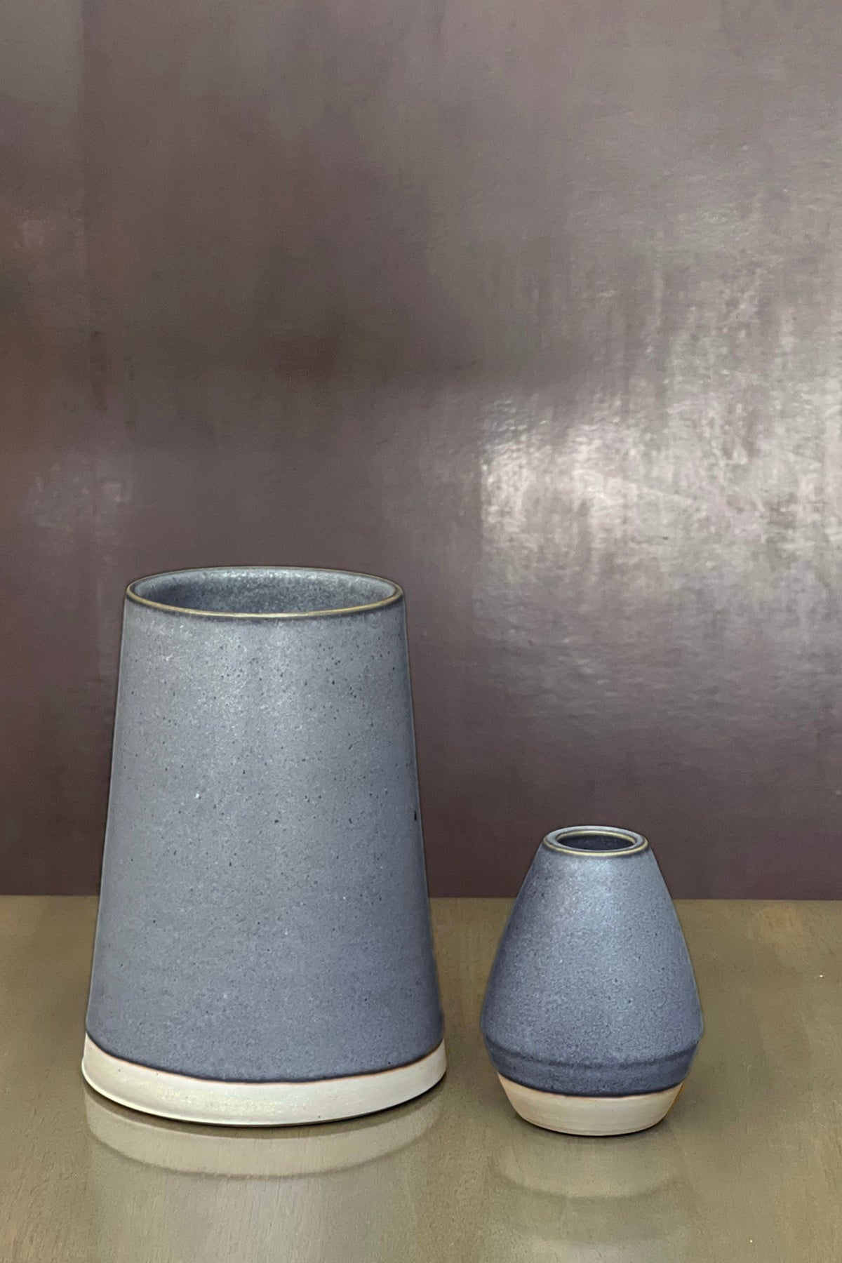   The CERAMIC LARGE VASE from Jenny Graham Home, featuring a stunning matte, blue-gray finish, is showcased alongside its smaller counterpart as an elegant centerpiece on a reflective surface against a neutral background. The casual design and earthy hues of these ceramic vases add charm to any setting. 